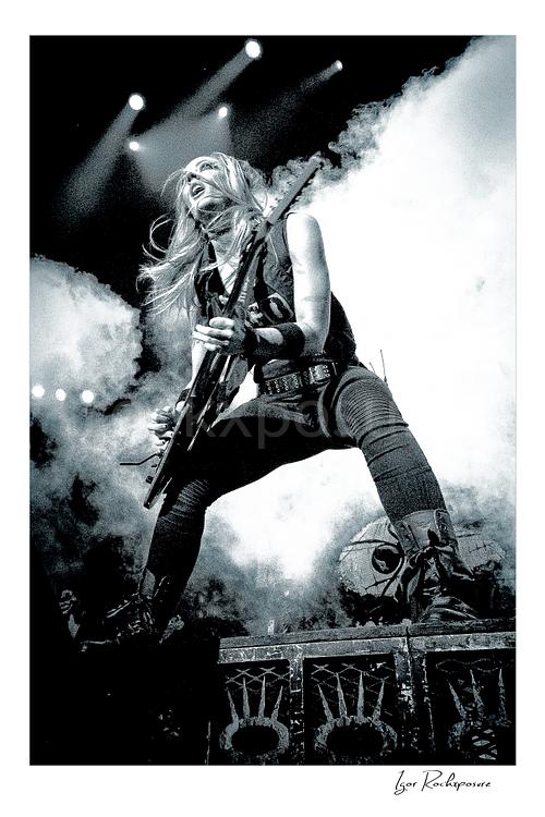 Vertical black and white image of Nita Strauss performing live on stage with an electric guitar, surrounded by thick stage smoke