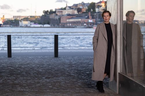 Helkroppsporträtt av kvinna i lång kappa vid glasfasad med reflektion, vatten och stadsmiljö i bakgrunden, Stockholm.