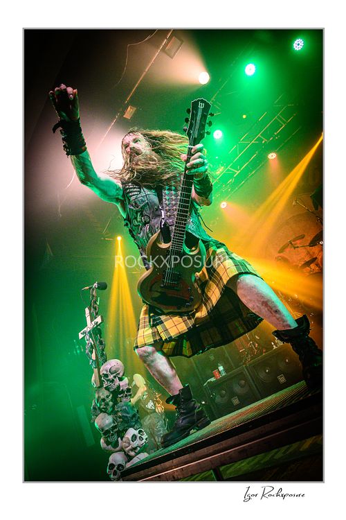 Low-angle vertical shot of Zakk Wylde standing on a skull-covered riser, wearing a kilt, holding his guitar with one hand and raising his other fist under green and yellow stage lights.