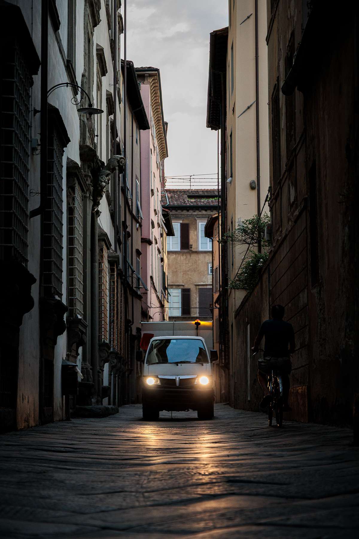 Twilight street scene in a narrow alley in Lucca, Italy, with a white van in the center of the cobblestone lane, headlights glowing warmly on the ground, a cyclist passing on the right in dark clothing, and tall beige, pink and brown buildings on both sides under an overcast evening sky, creating a calm, moody atmosphere.