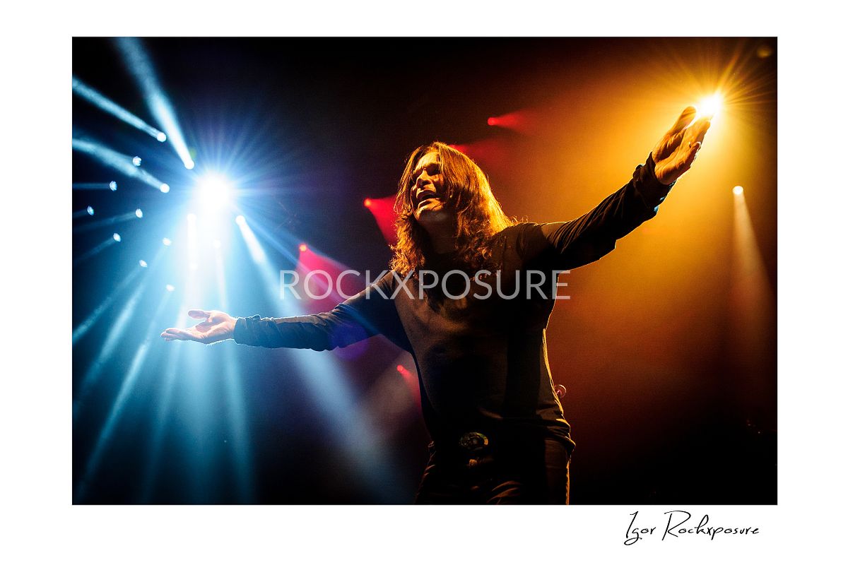 Ozzy Osbourne of Black Sabbath standing with arms raised wide under dramatic blue and orange stage lighting during a live performance