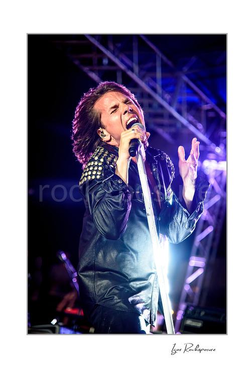 Vertical color image of Joey Tempest of Europe performing live on stage with a white microphone stand, black leather jacket, bright backlight, and expressive singing pose