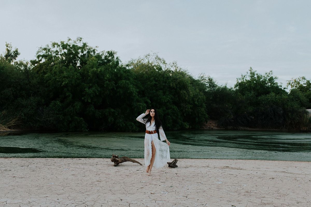 Ethereal boudoir portrait of a woman in a flowing white lace gown walking gracefully with vibrant blue water in the background, blending elegance with natural serenity.