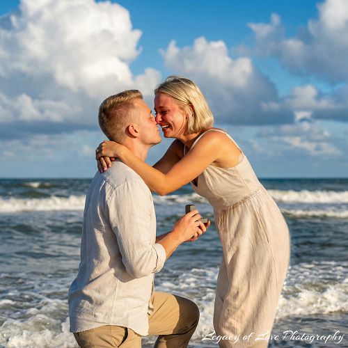 A man on one knee proposals to a surprised woman with a ring box on a Jacksonville Beach during the golden hour sunset. A passionate kiss is shared with the ocean and clouds behind them.