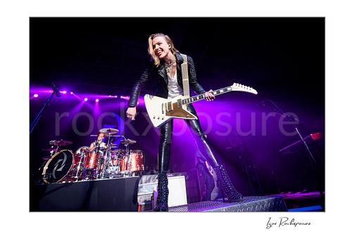 Horizontal color image of Lzzy Hale of Halestorm performing live with a white guitar at the front of the stage, smiling under vivid purple concert light with Arejay Hale visible at the drum kit behind her