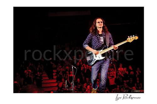 Horizontal color image of Geddy Lee performing with his bass guitar during a Rush concert, walking across the stage with red-lit audience in the background