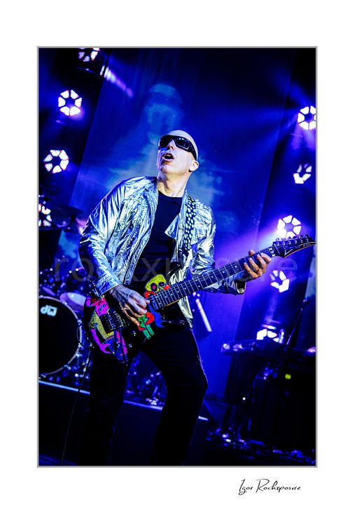 Vertical color image of Joe Satriani performing live in silver jacket and sunglasses with decorated black guitar under blue stage lights