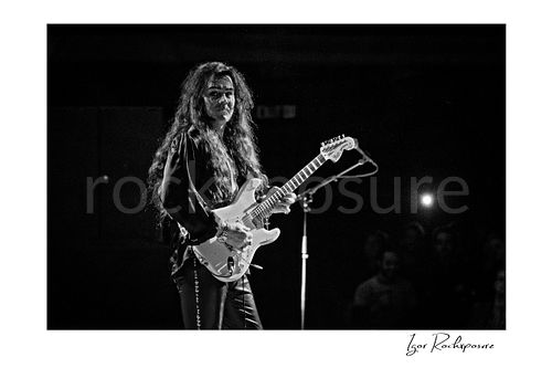 Horizontal black and white image of Yngwie Malmsteen performing live with a white guitar, looking toward the camera with long hair flowing and the stage microphone and audience lights visible