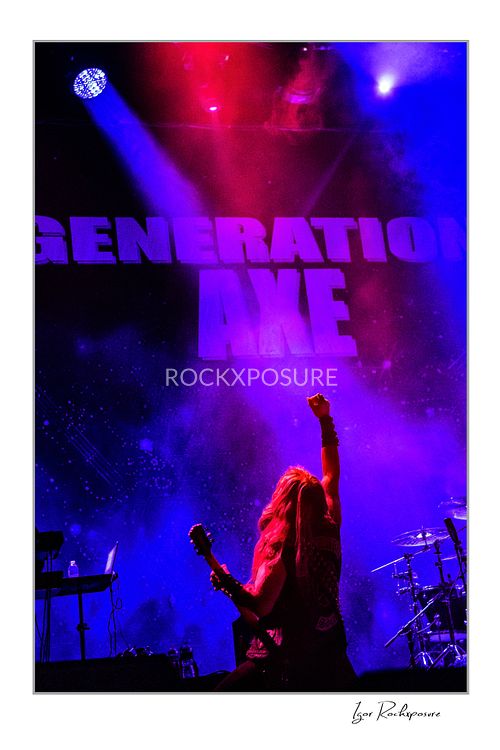 Shot from behind of Zakk Wylde on stage with his guitar, raising his right fist toward a large Generation Axe backdrop under vibrant purple and red lighting.
