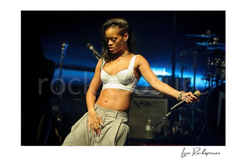 Horizontal color image of Rihanna performing on stage in a white top with a gold microphone and deep blue concert lighting