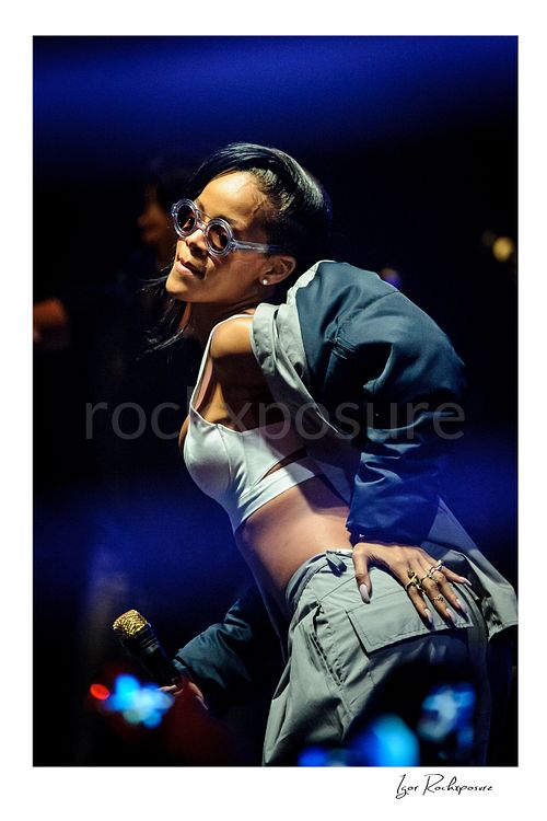 Vertical color image of Rihanna posing on stage in a white top and draped jacket with a gold microphone and deep blue concert lighting