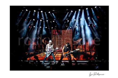 Horizontal color image of Rudolf Schenker and Paweł Mąciwoda of Scorpions performing live under dramatic white spotlights with glowing orange stage screens