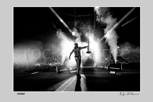 Horizontal black and white image of Bret Michaels on a Poison stage, captured with a remote camera mounted on the drum kit facing forward, with smoke blasts and bright backlight behind him