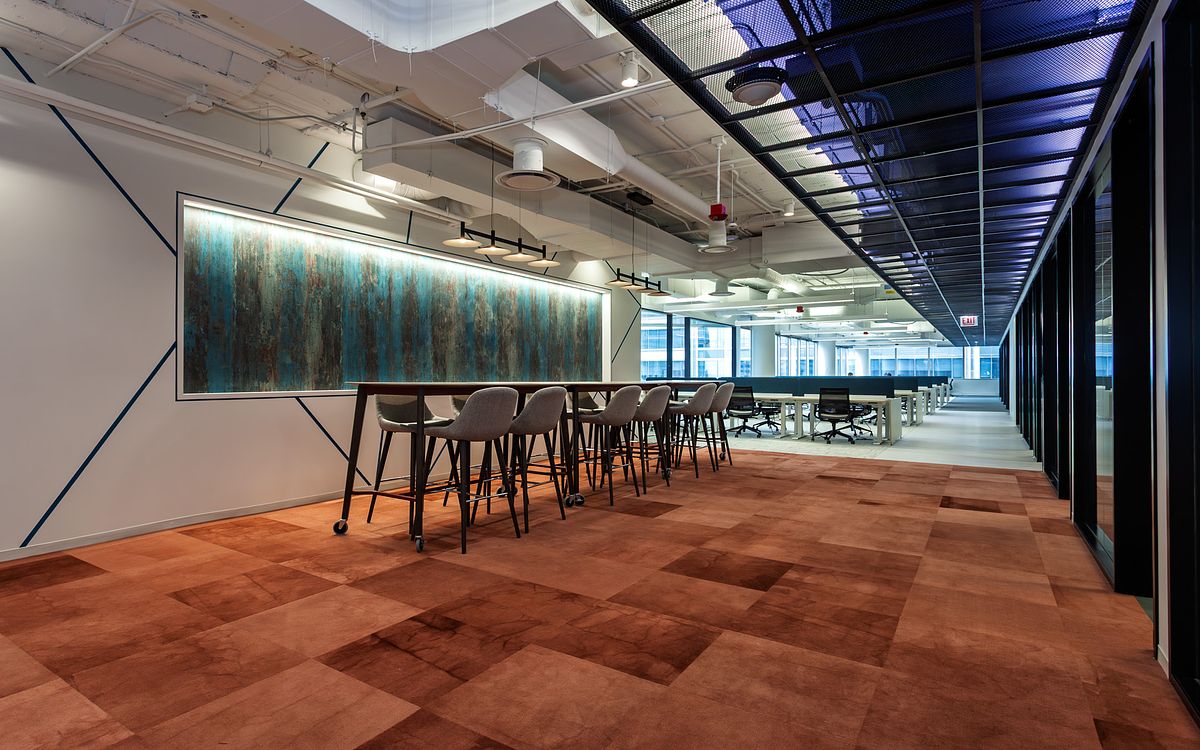 A modern Chicago office communal area at 1 South Wacker featuring a long high-top work table with grey stools, a large teal and gold abstract mural, and orange-toned carpet tiles.