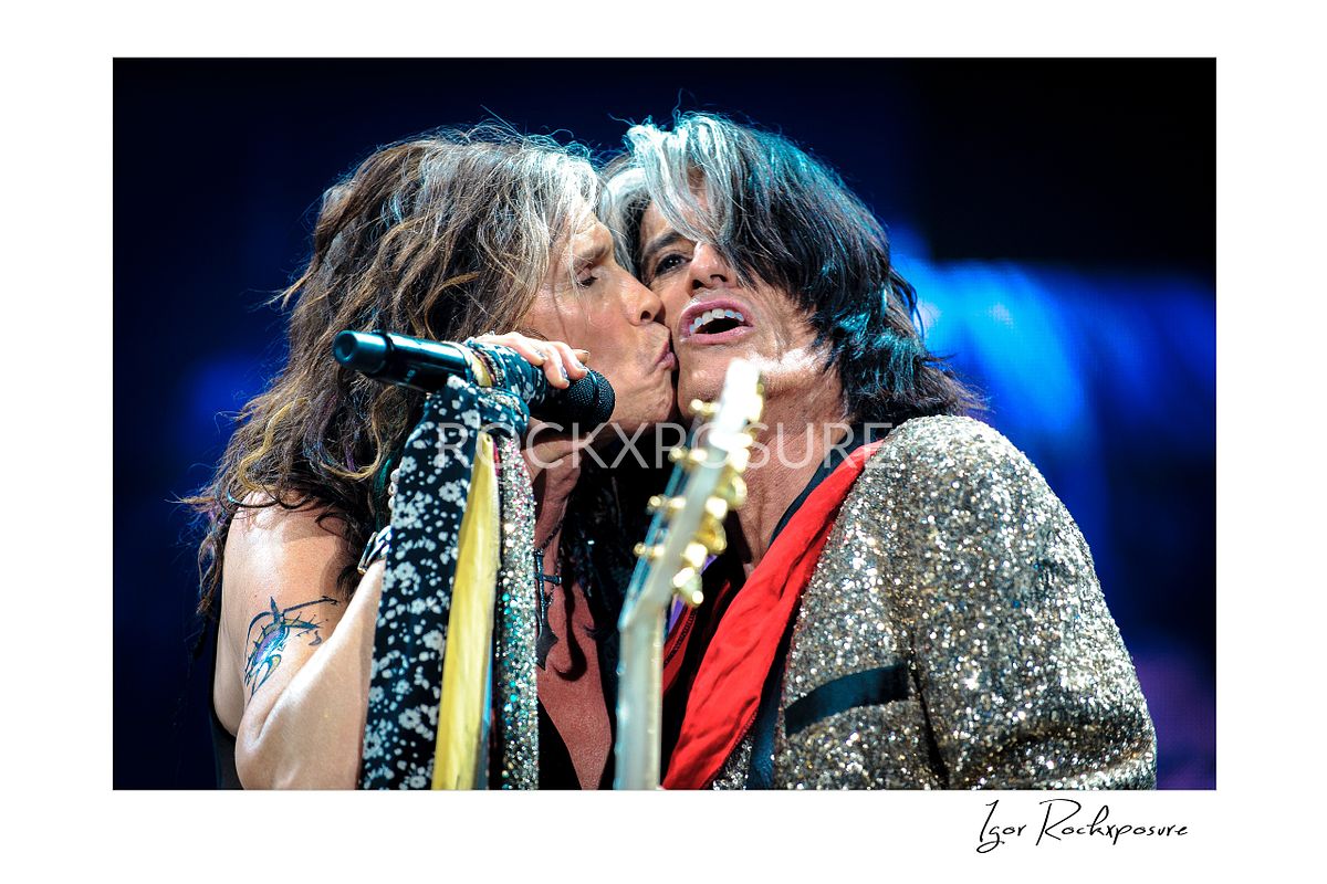 Horizontal concert photography of Steven Tyler kissing Joe Perry on the cheek at the microphone during a live Aerosmith performance under blue stage lighting