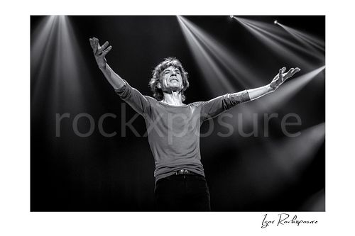 Horizontal black and white image of Mick Jagger of The Rolling Stones standing with both arms outstretched beneath dramatic stage spotlights