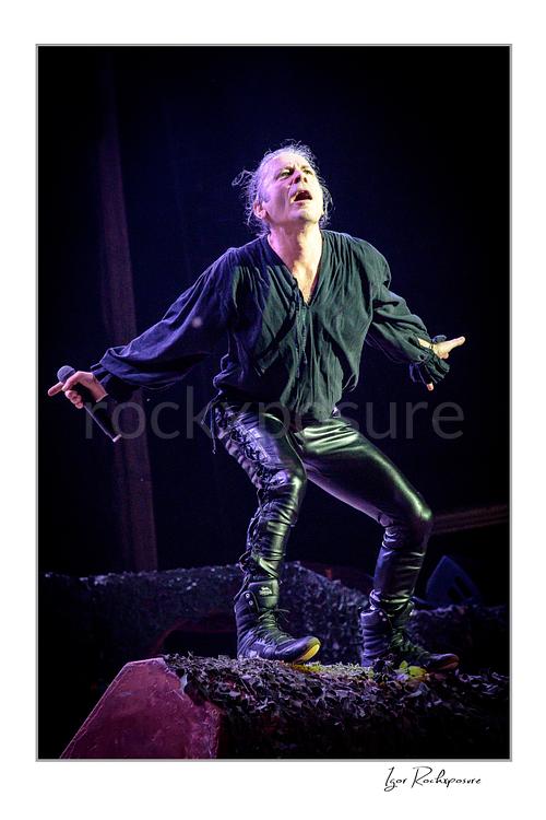 Vertical color image of Bruce Dickinson of Iron Maiden performing live on stage, holding a microphone and striking an expressive pose under concert lighting.