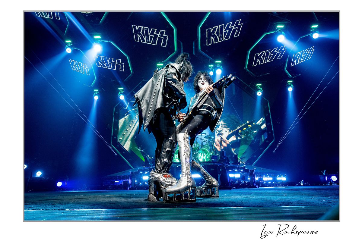 Gene Simmons and Tommy Thayer of KISS performing together in a wide stage stance with bass and guitar under bright blue arena stage lighting with band stage set behind them