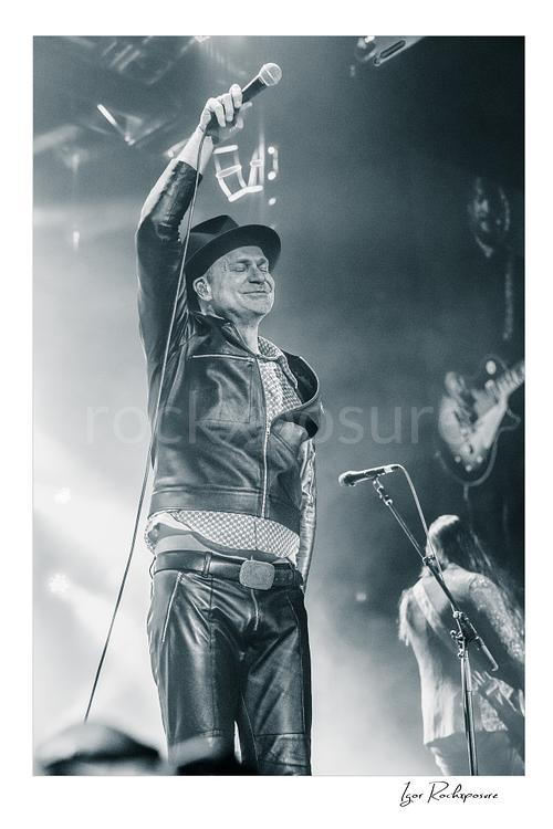 Vertical black and white image of Gord Downie of The Tragically Hip raising a microphone high above his head, wearing a hat and leather jacket in bright concert haze with a blurred band member behind him