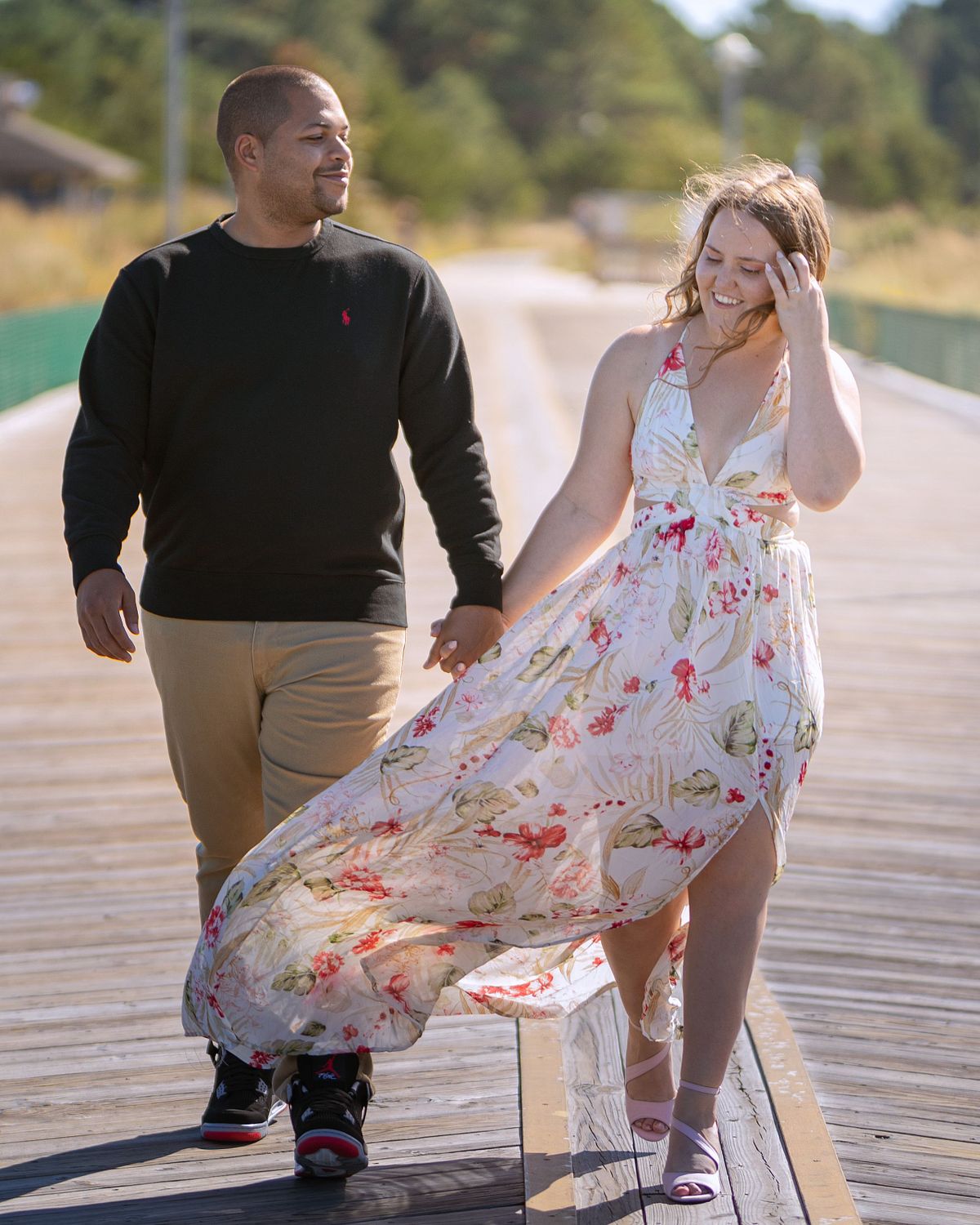 couple walking on the pier at cape henlopen, sussex county, de, the woman is wearing a long, flowery dress, the man is wearing a black shirt and tan pants, they are holding hands and smiling at each other