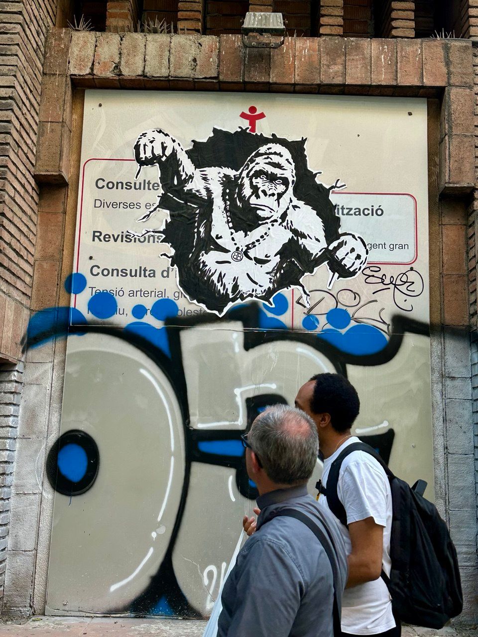 Street art stencil in Barcelona depicting a powerful gorilla appearing to burst through a sign, wearing a necklace with the prominent Street Cyber symbol. This piece symbolizes strength, primal power, and the disruptive force of the Street Cyber movement, subtly integrating brand identity with a universally recognized image of raw energy. It is set against a backdrop of blue graffiti.