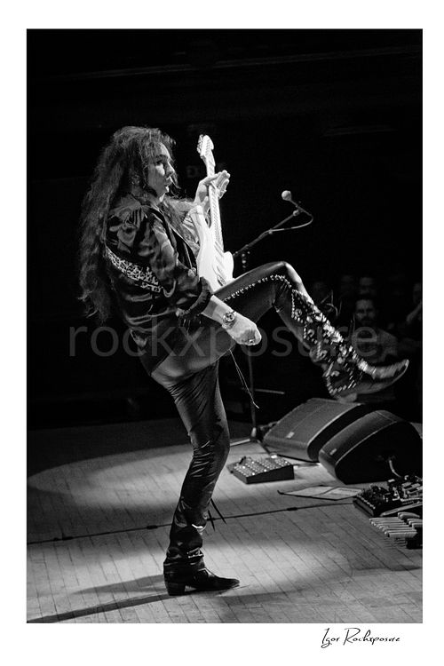 Vertical black and white image of Yngwie Malmsteen performing live with a white guitar, lifting one leg high in a dramatic stage pose beside the microphone and floor monitors