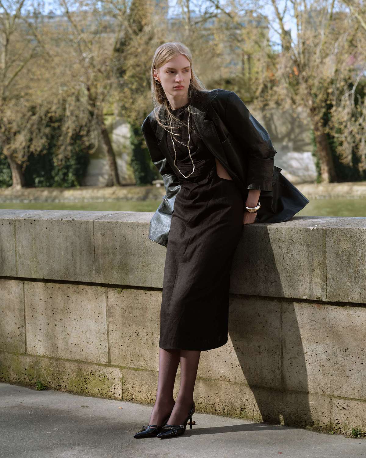 A full frame shot of a woman in a black leather jacket, black dress, and heels leans against a wall