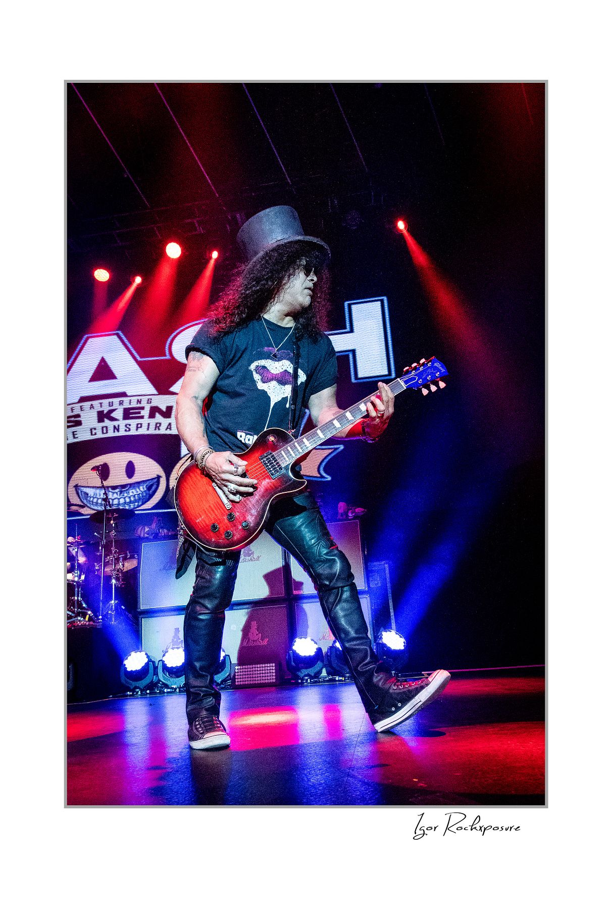 Vertical concert photography of Slash playing electric guitar in a wide stance under red and blue stage lighting