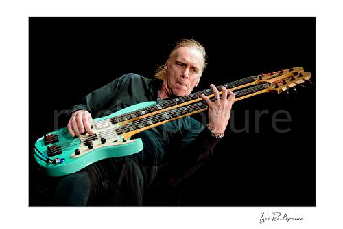 Horizontal color image of Billy Sheehan playing his double-neck bass in a tight low-angle live performance shot