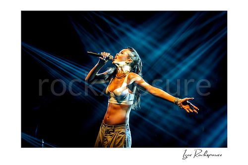 Horizontal color image of Rihanna singing into a microphone with one arm outstretched beneath dramatic blue stage beams