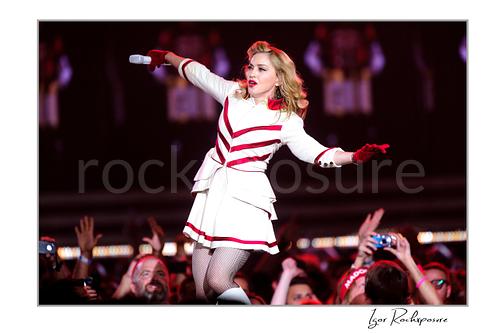 Horizontal color image of Madonna performing in a white and red stage outfit while moving through the crowd with a microphone in hand
