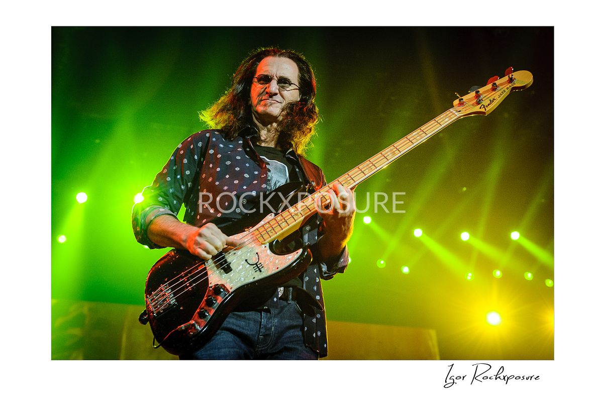 Horizontal concert photography of Geddy Lee playing bass guitar under green stage lighting