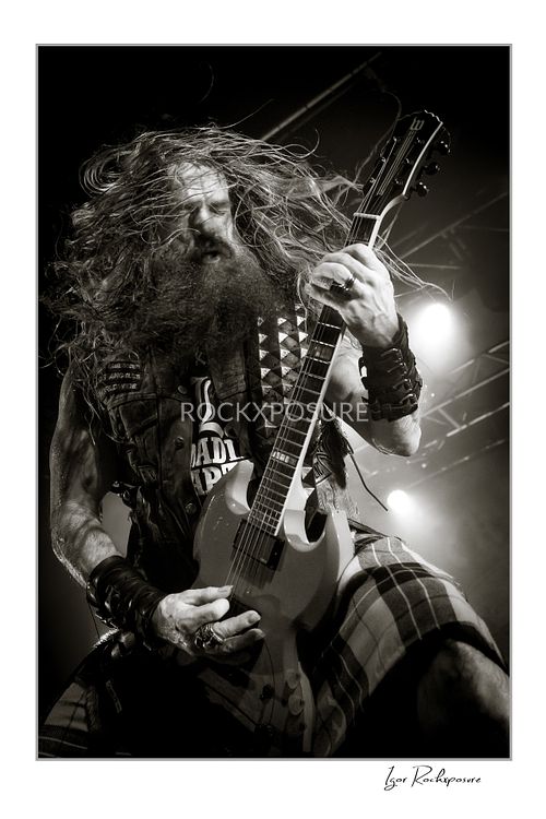 Black and white vertical shot of Zakk Wylde playing guitar on stage, with his long hair flying and wearing a plaid kilt