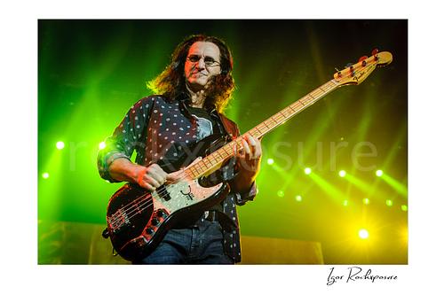 Horizontal color image of Geddy Lee performing with his bass guitar during a Rush concert under vivid green and yellow stage lights
