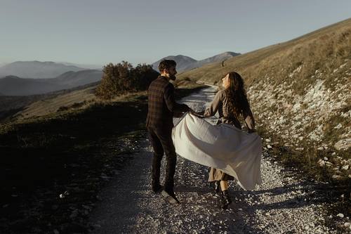 Donna che corre sollevando il vestito su strada di montagna, seguita dal compagno, scena dinamica al tramonto.