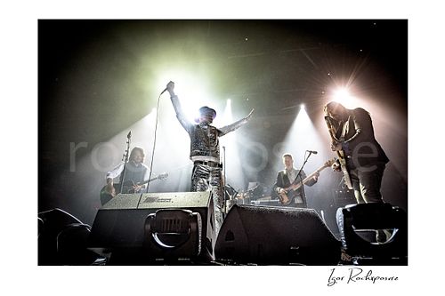 Horizontal color image of The Tragically Hip performing live on stage, with Gord Downie in a glittering silver suit at center with arms raised high while band members play under bright concert spotlights