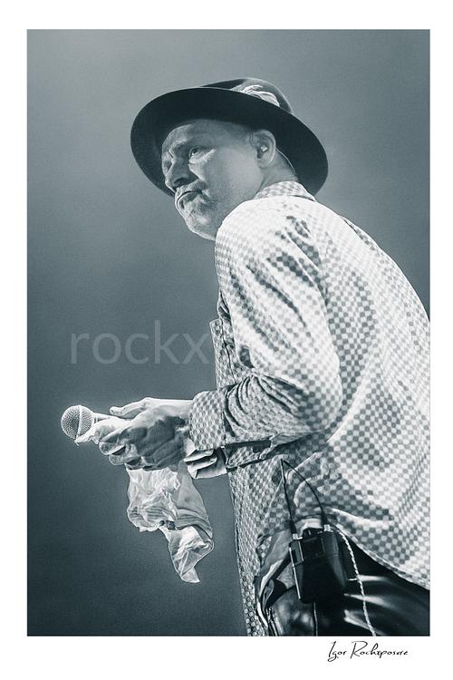 Vertical black and white image of Gord Downie of The Tragically Hip holding a microphone and white cloth in a quiet live performance moment, wearing a hat and patterned shirt under soft stage light