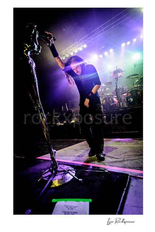 Vertical color image of Korn singer Jonathan Davis standing on stage beside his distinctive stage microphone stand with the drum kit visible behind him