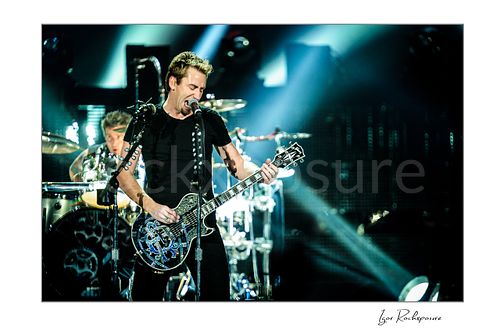 Horizontal color image of Chad Kroeger of Nickelback singing into a microphone while playing guitar with the drummer visible behind him under bright concert beams