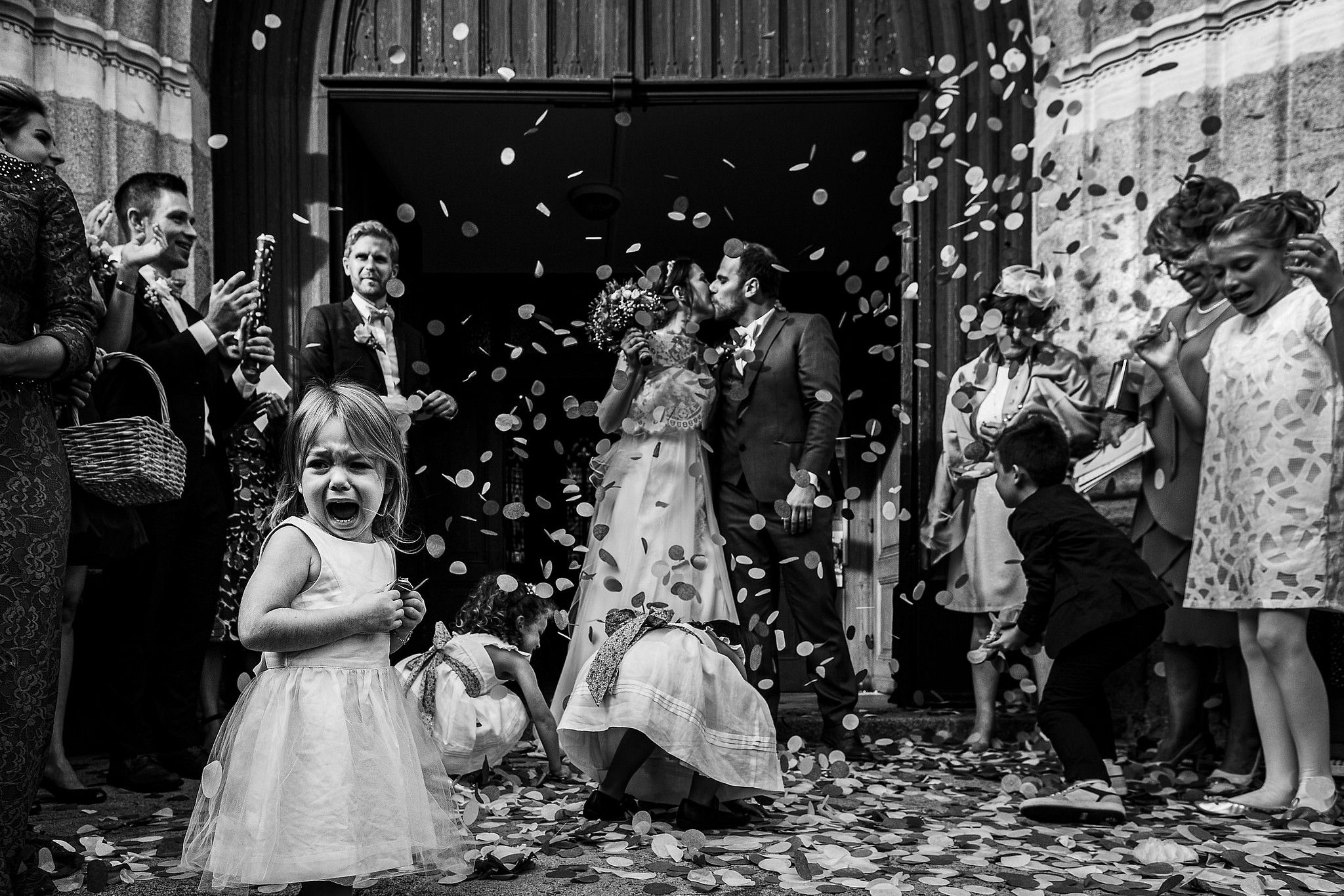 Enfant qui pleure &agrave; la sortie de l'&eacute;glise devant le couple des mari&eacute;s qui s'embrassent captur&eacute; par S&eacute;bastien CLAVEL photographe de Mariage &agrave; Lyon et Gen&egrave;ve