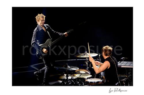 Horizontal color image of Matt Bellamy and Dominic Howard of Muse performing live on stage with guitar and drums against a dark concert background