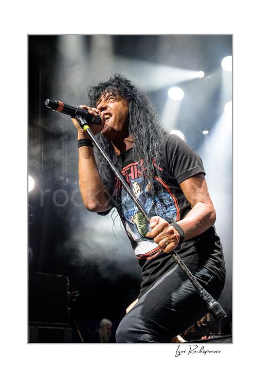 Vertical color image of Joey Belladonna of Anthrax performing live on stage, singing into a microphone under bright concert lights