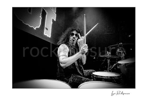 Horizontal black and white image of Frankie Banali at the drum kit holding drumsticks and raising his middle finger in a tight live performance shot
