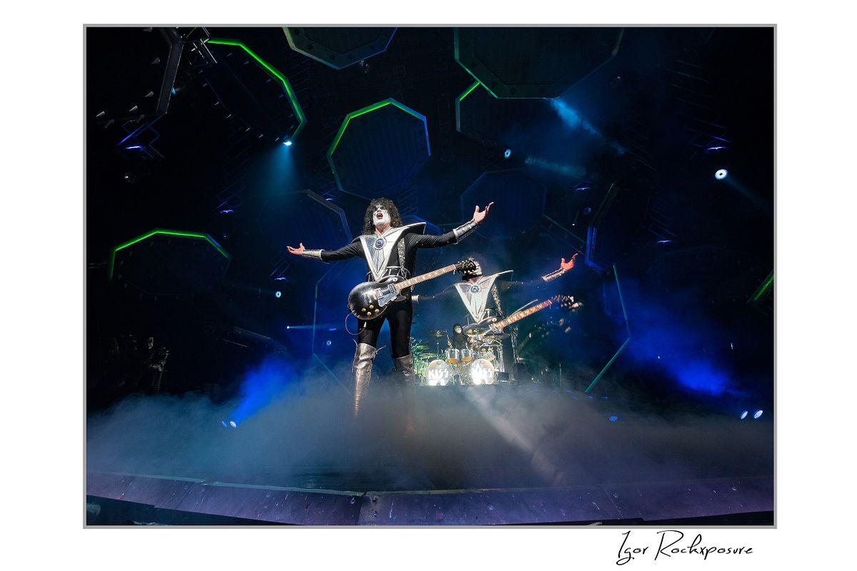 Tommy Thayer of KISS performing in a wide guitar stance under blue arena stage lighting with illuminated stage arches behind him