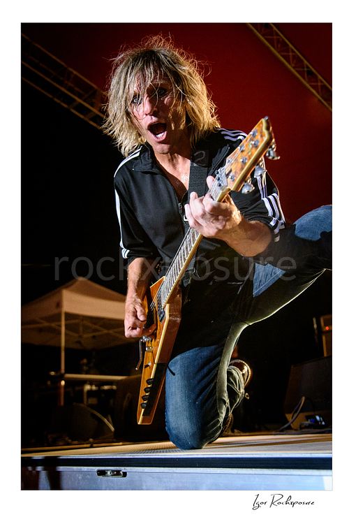 Vertical color image of C.C. DeVille performing with electric guitar during a Poison concert, kneeling at the front of the stage under vivid red light