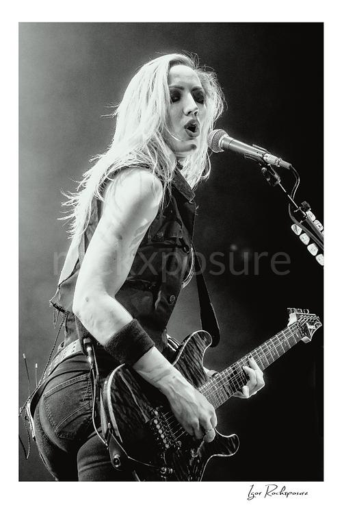 Vertical black and white image of Nita Strauss performing live on stage with an electric guitar beside a microphone