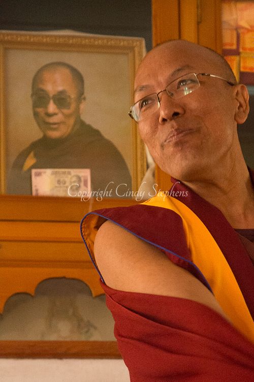 A smiling Buddhist monk in traditional robes stands before a portrait of the current Dalai Lama, embodying peace and spirituality.