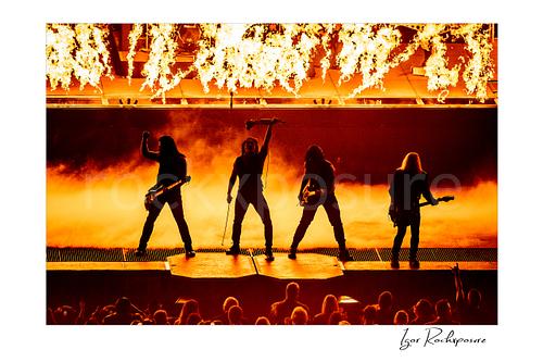 Horizontal color image of Trans Siberian Orchestra performing in silhouette with guitars, raised microphone stand, flames, and stage smoke