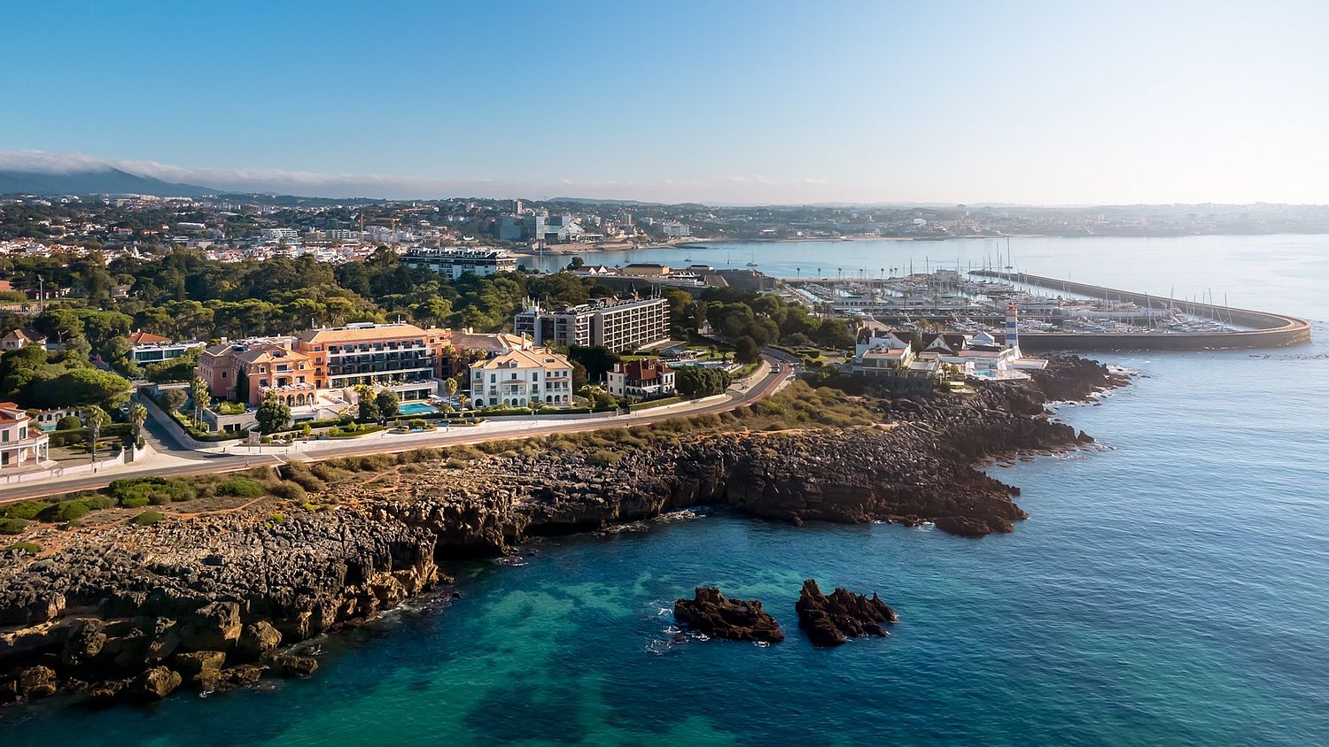 Vista aérea do Grande Real Villa Itália Hotel & Spa em Cascais, membro dos Leading Hotels of the World, com a marina e o farol de Santa Marta ao fundo, fotografada por Pedro Brás Photography — fotografia aérea profissional de hotelaria e resorts de luxo e