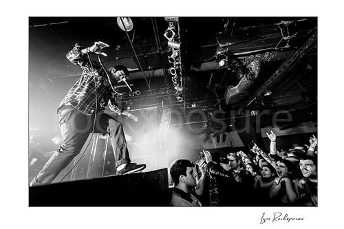 Horizontal black and white image of Chino Moreno of Deftones performing live on stage above the crowd with a handheld microphone, outstretched arm, and fans reaching up